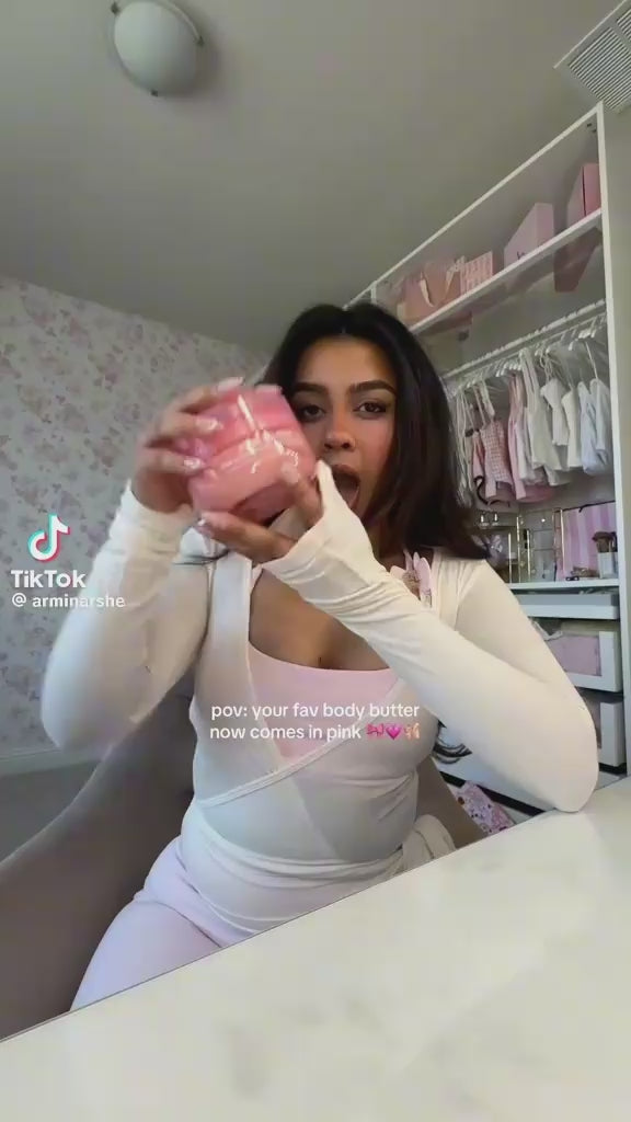 Woman holding pink body butter container displaying skincare product in bedroom